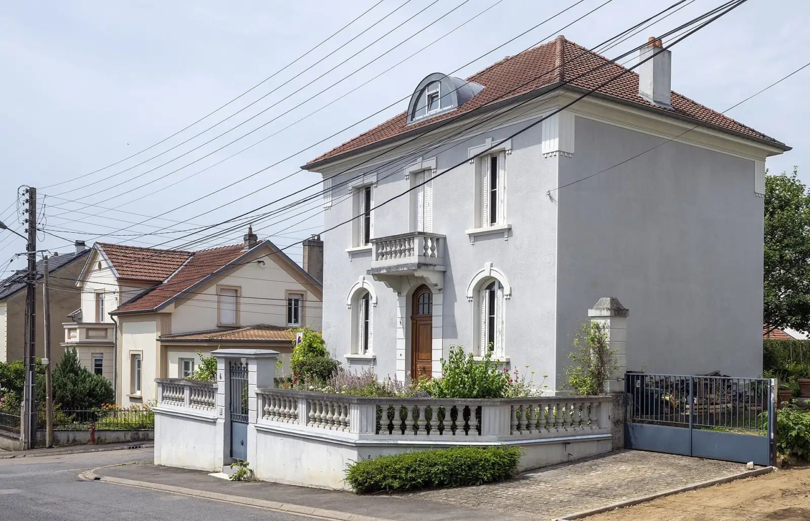 Maison individuelle crépie de gris, avec balcon et muret à balustres, au coin d'une rue résidentielle de Hettange-Grande