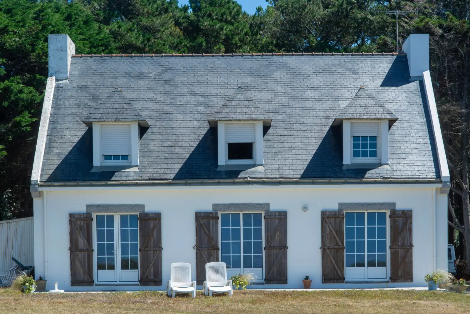 Maison blanche à toit d'ardoise avec lucarnes, volets en bois et petit jardin devant la façade