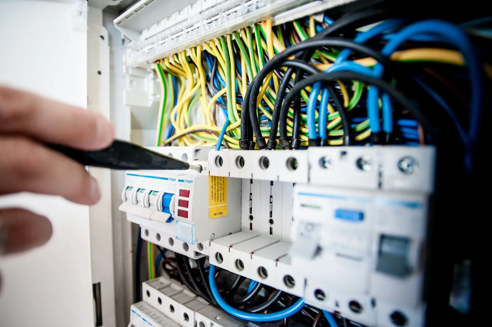 Main d'un électricien pointant un tableau électrique ouvert, disjoncteurs et câbles de couleur apparents