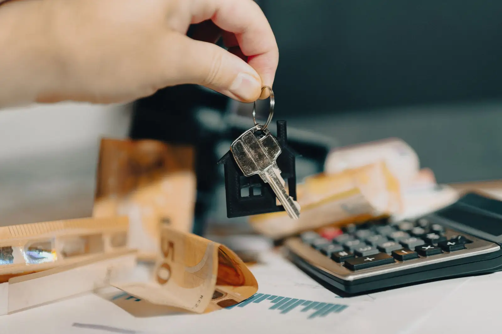 Main tenant un trousseau de clés au-dessus de billets en euros et d'une calculatrice posés sur un bureau