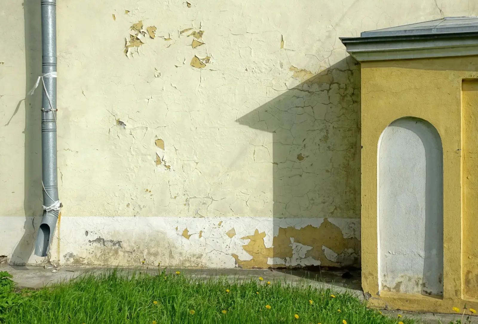 Pied d'un vieux mur dont l'enduit s'écaille, près d'une descente d'eaux pluviales et d'une pelouse