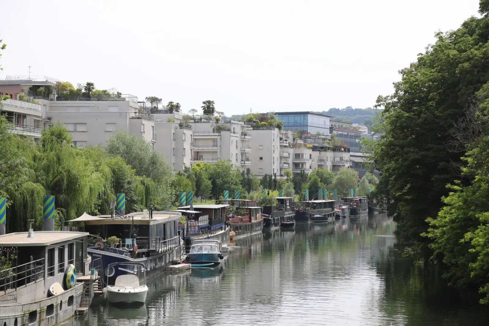 Péniches et bateaux-logements amarrés le long d'un canal bordé d'arbres, avec des immeubles en arrière-plan