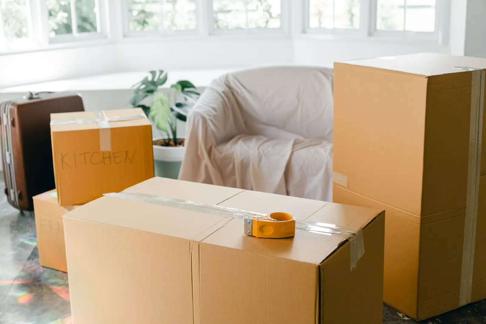Cartons de déménagement fermés au ruban adhésif, valise et fauteuil couvert d'un drap dans une pièce lumineuse