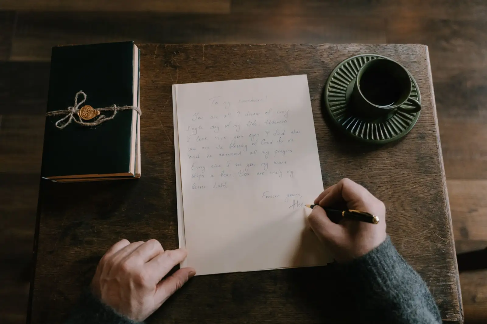Personne rédigeant une lettre à la main sur une table en bois, à côté d'un carnet et d'une tasse de café