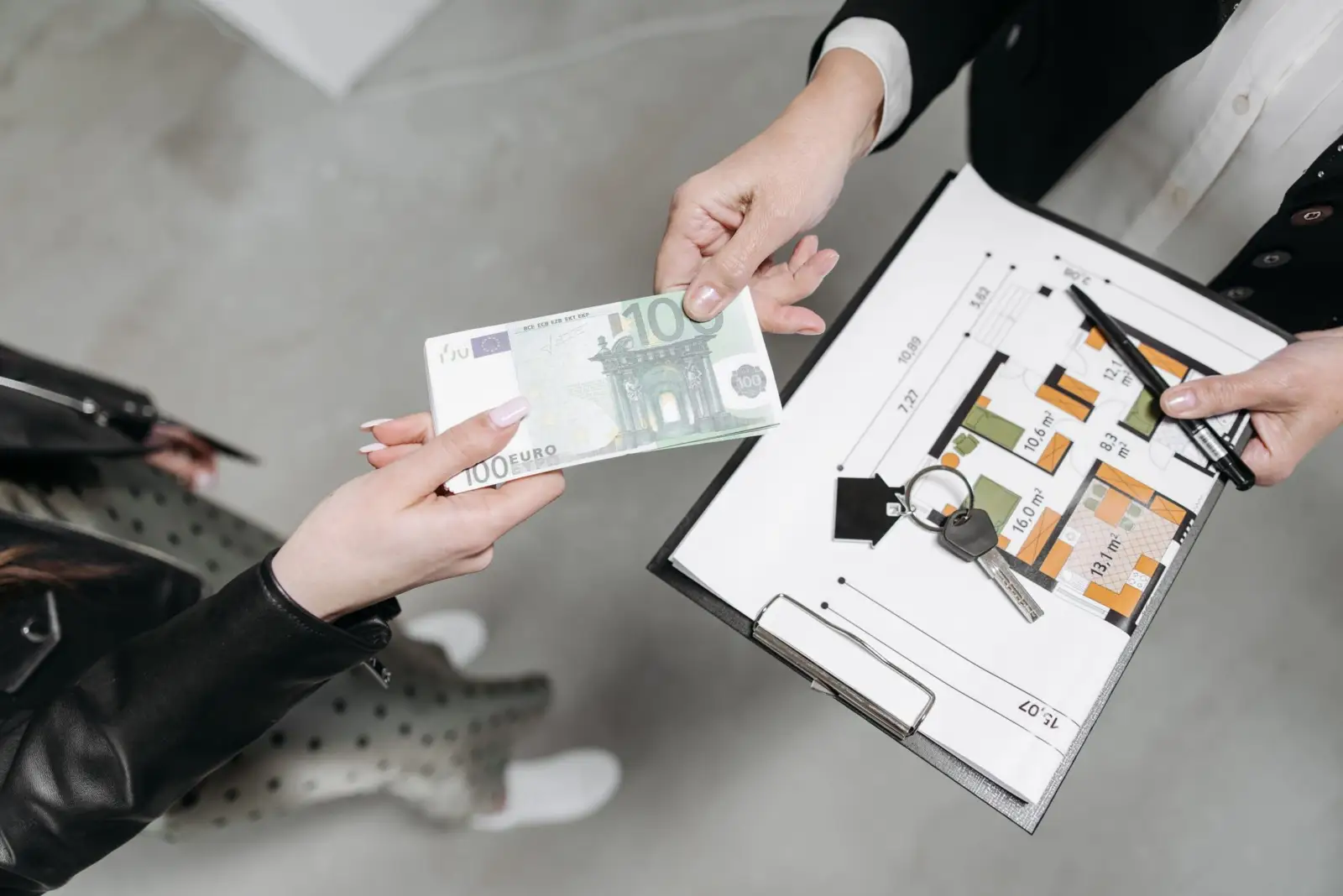 Deux personnes échangent un billet de 100 euros et un trousseau de clés au-dessus d'un plan d'appartement posé sur un porte-bloc