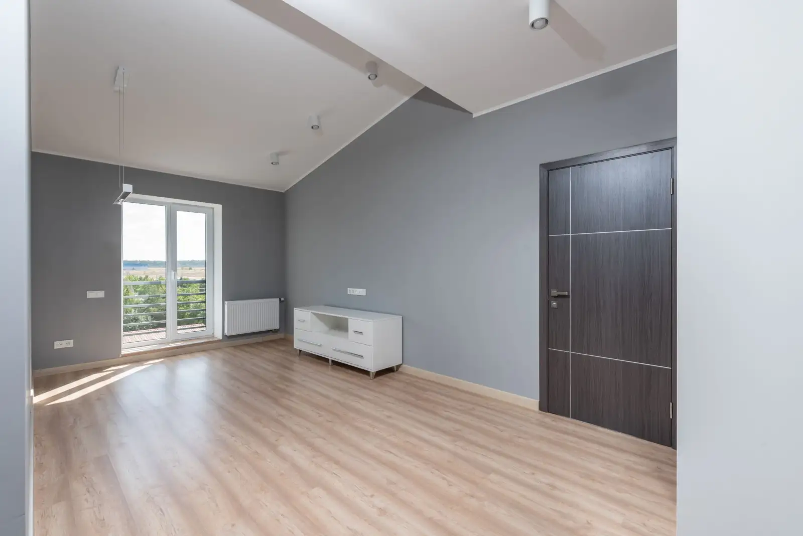 Pièce vide aux murs gris avec parquet clair, radiateur sous une porte-fenêtre et meuble bas blanc
